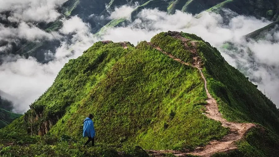 A trekker in a rain poncho walking the Tà Xùa ridge with cloud pouring through the valleys below